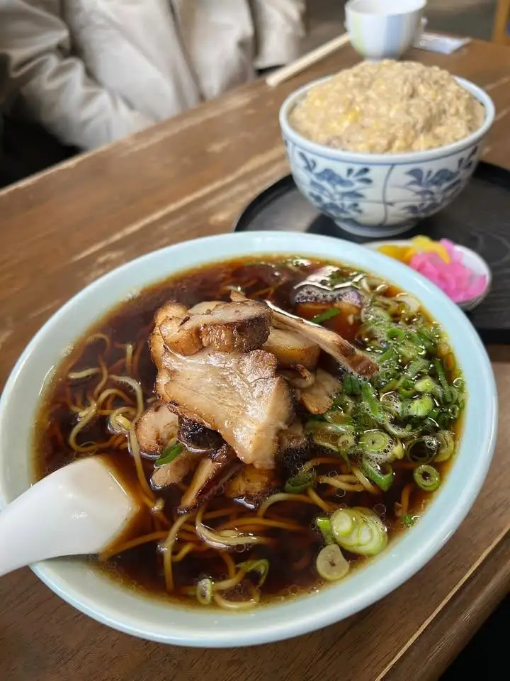 a close-up photo of a steaming bowl of ramen with chashu pork, green onions, and broth, placed on a wooden table next to a…