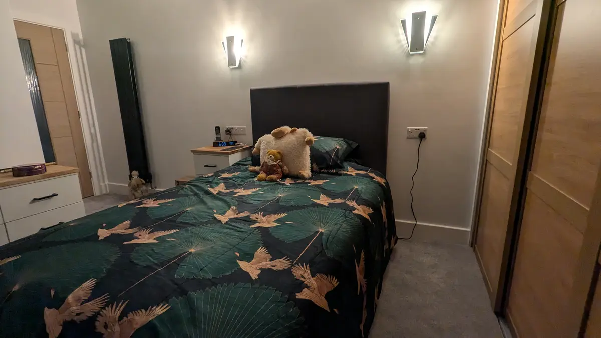 A cozy bedroom with a bed covered in a dark green duvet featuring large lily pads and golden birds. Two stuffed animals, a…