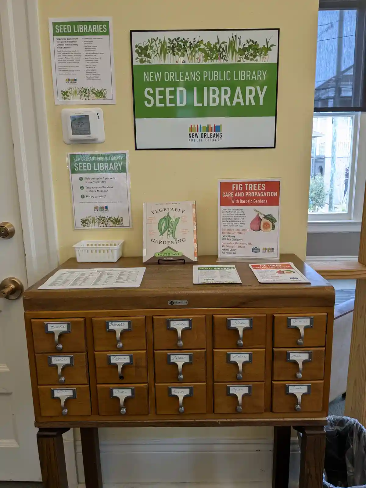 A wooden cabinet with labeled drawers for seeds like 'Beets', 'Broccoli', and 'Tomatoes' sits in a library, under signs for…