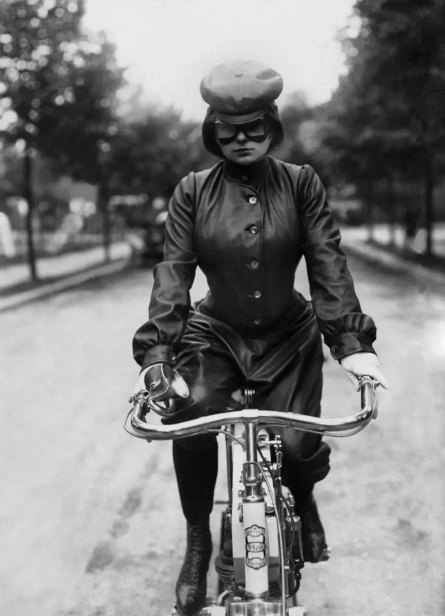 A vintage black-and-white photo shows a woman in full leather cycling gear, including a cap, goggles, gloves, and boots…