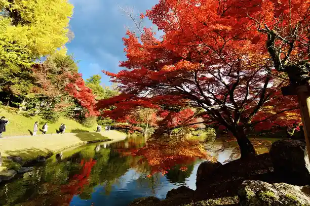 A scenic autumn photo shows vibrant red and yellow maple trees reflecting in a calm pond, with people walking along a grassy…