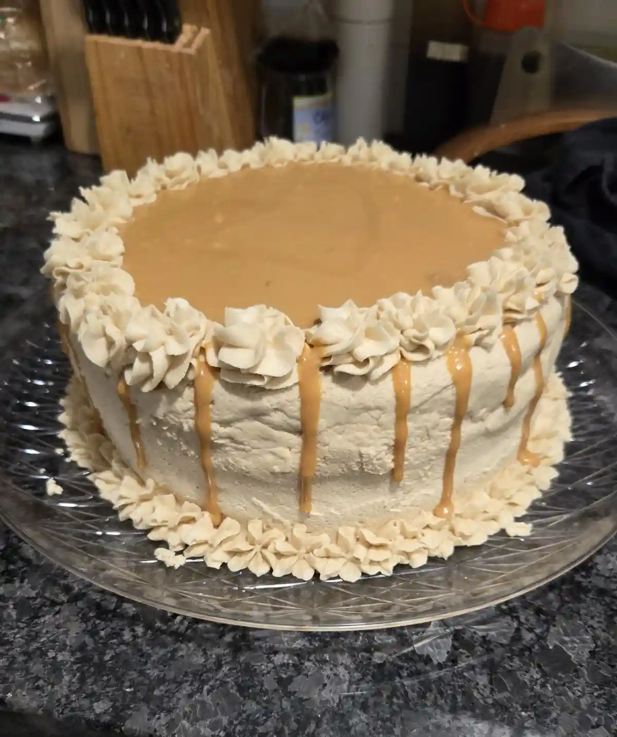 A round cake with light-colored frosting, topped with caramel drizzle and piped frosting flowers around the top and bottom…