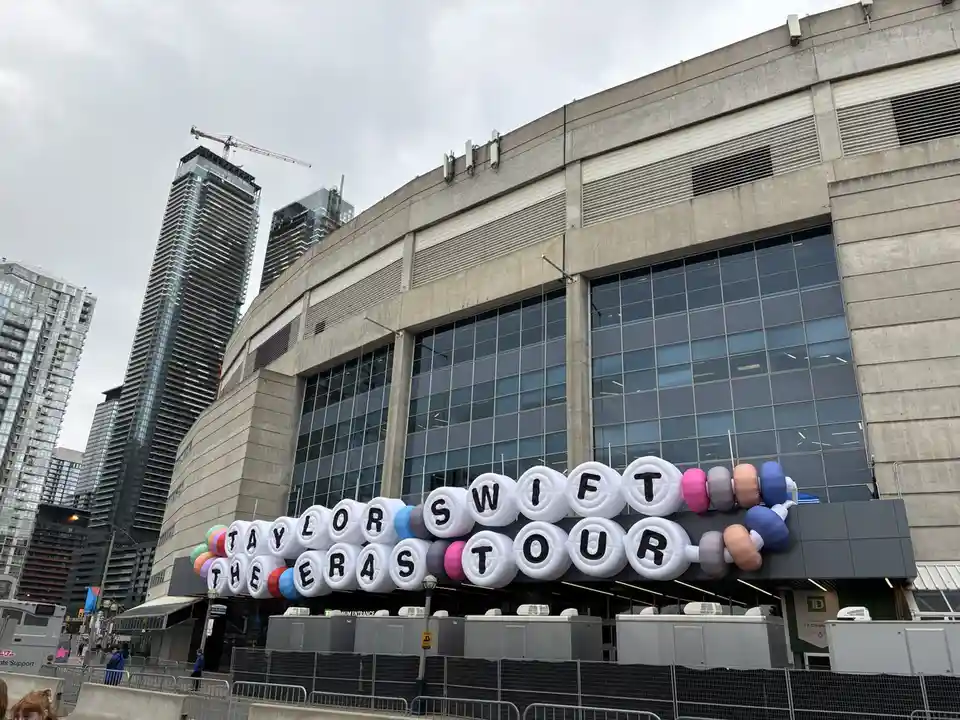 An outdoor photo of a large stadium with a massive inflatable sign spelling out 'TAYLOR SWIFT THE ERAS TOUR' in large white…