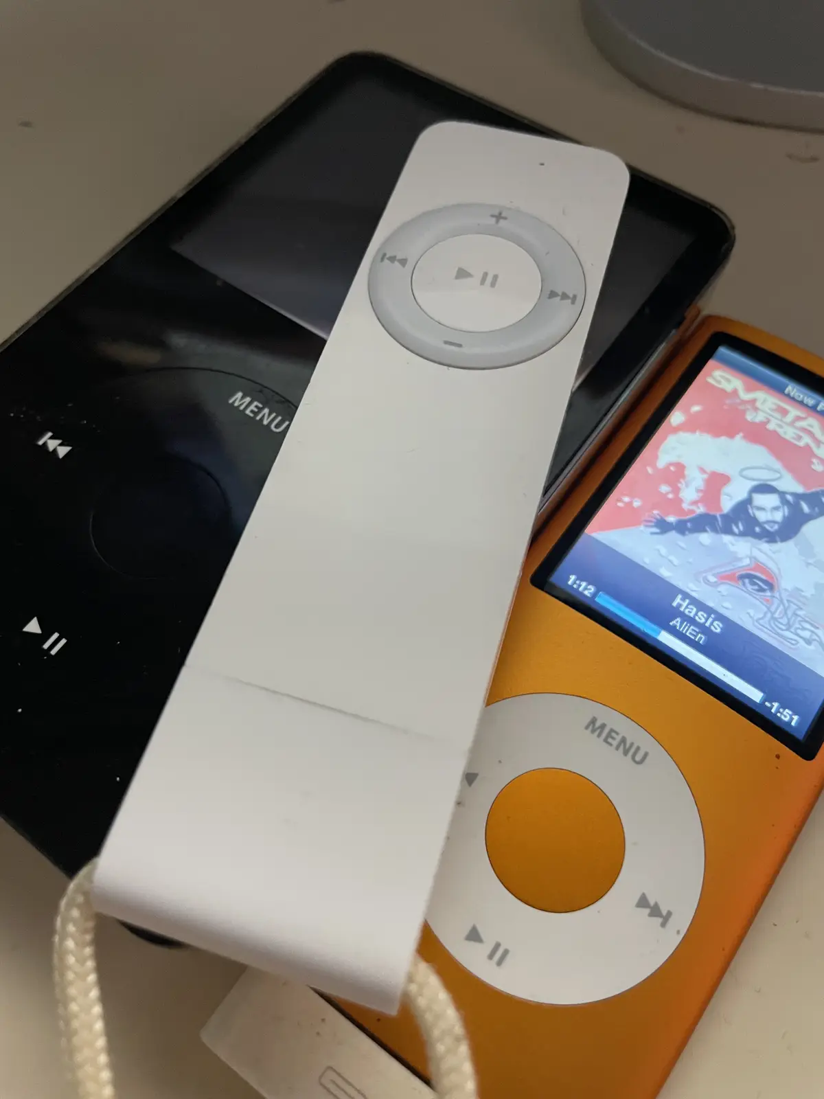 A white iPod shuffle is placed on top of a black and an orange iPod classic, both showing their iconic click wheel…
