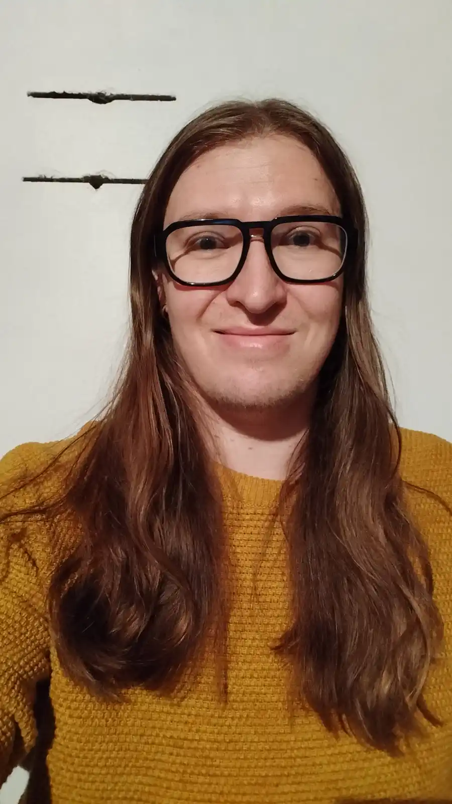 A person with long brown hair and black-rimmed glasses, wearing a mustard-yellow sweater, smiles at the camera while…