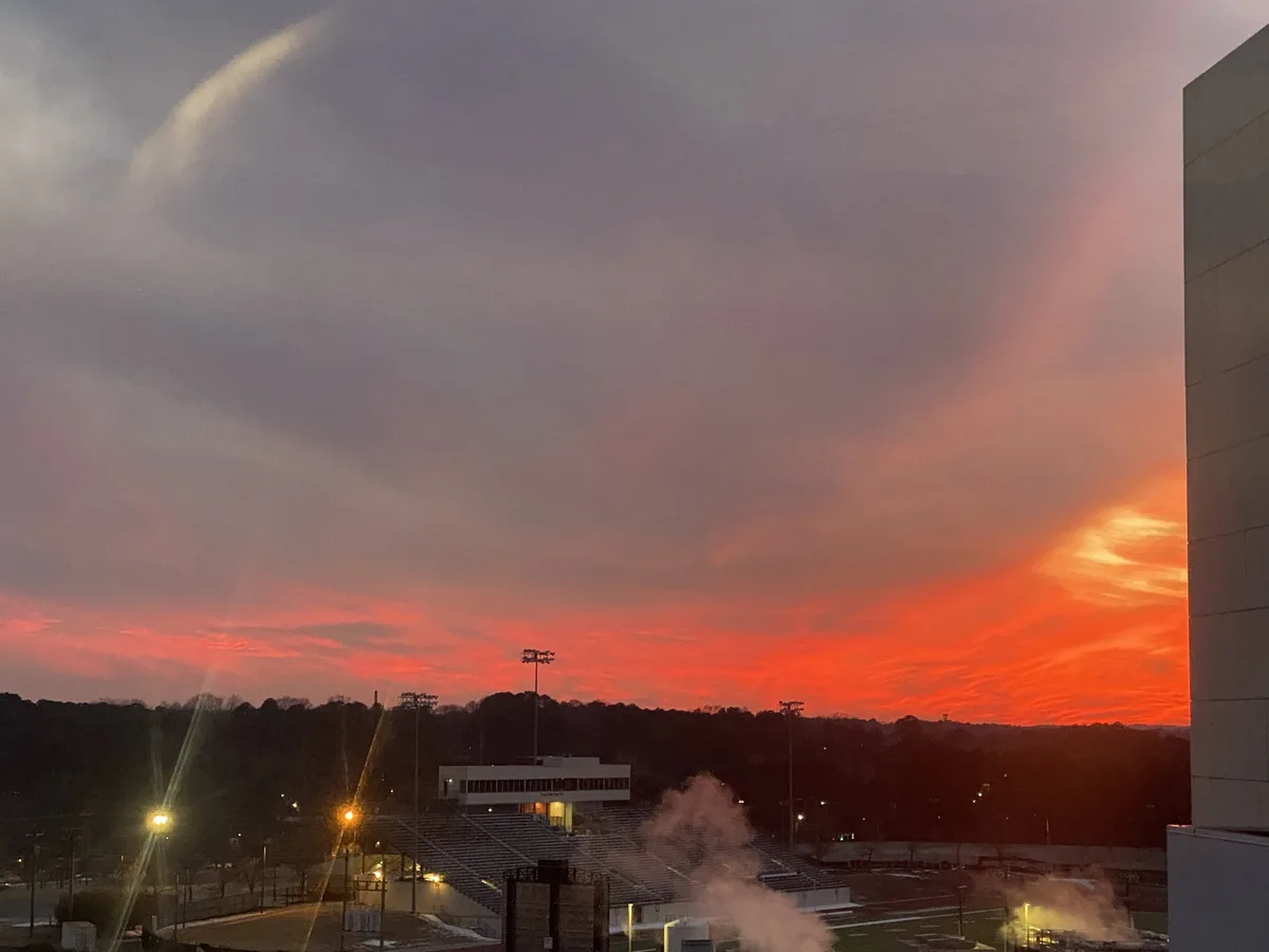 A dramatic sunset over a stadium with fiery orange and red clouds, stadium lights on, and steam rising from the field. The…