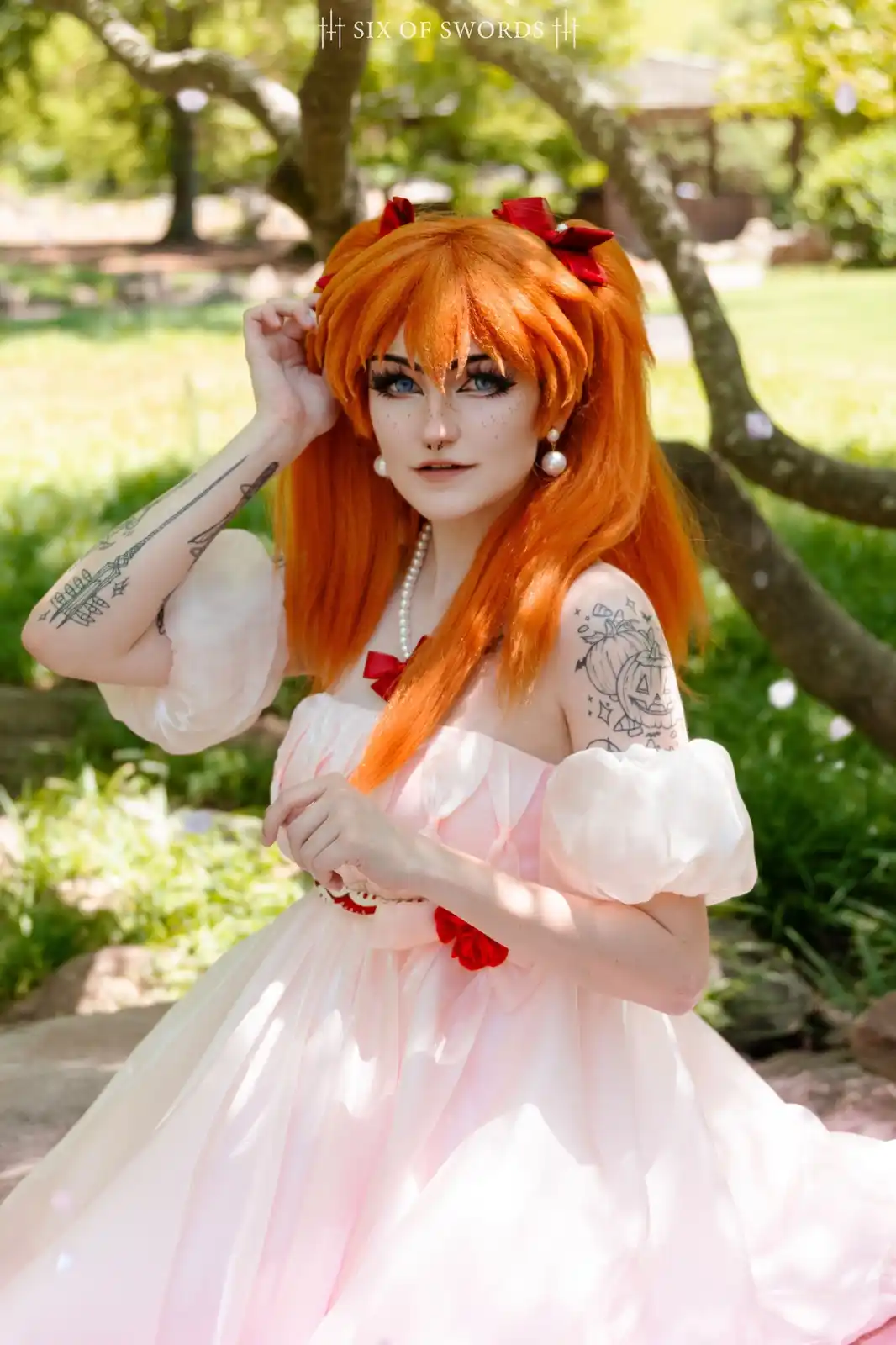 A person with bright orange hair styled in pigtails with red bows poses outdoors in a light pink off-the-shoulder dress with…