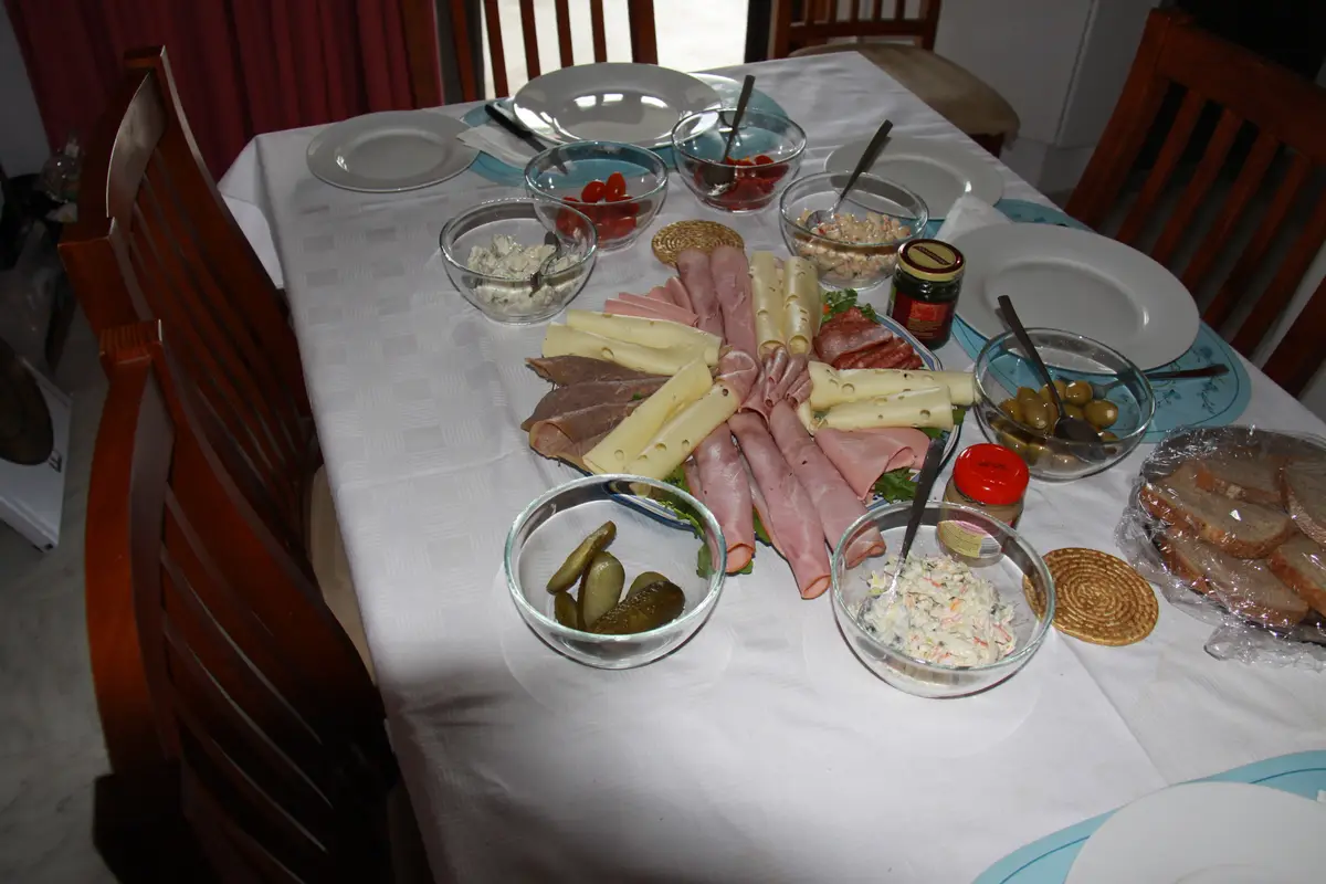 A dining table is set with a large charcuterie spread including sliced meats, cheese, pickles, olives, and various salads in…