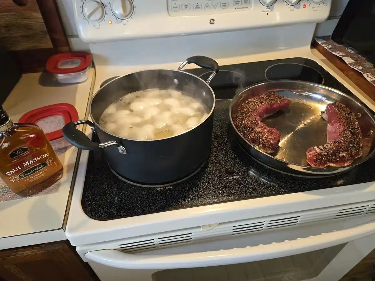 A white electric stove with a black glass cooktop has a pot of boiling water with dumplings and a pan with two raw, seasoned…
