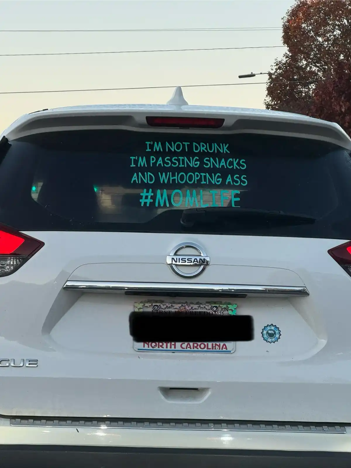 The rear of a white Nissan Rogue with a North Carolina license plate shows a teal decal on the back window that reads 'I'M…