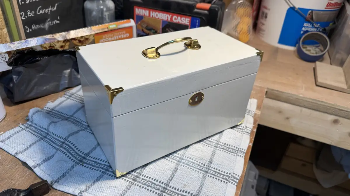 A white painted wooden box with brass hardware sits on a blue and white checkered cloth on a cluttered workbench. The box…