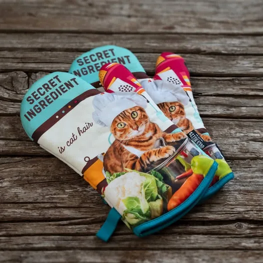 A pair of colorful oven mitts featuring a cartoon cat wearing a chef's hat, surrounded by vegetables like carrots and…