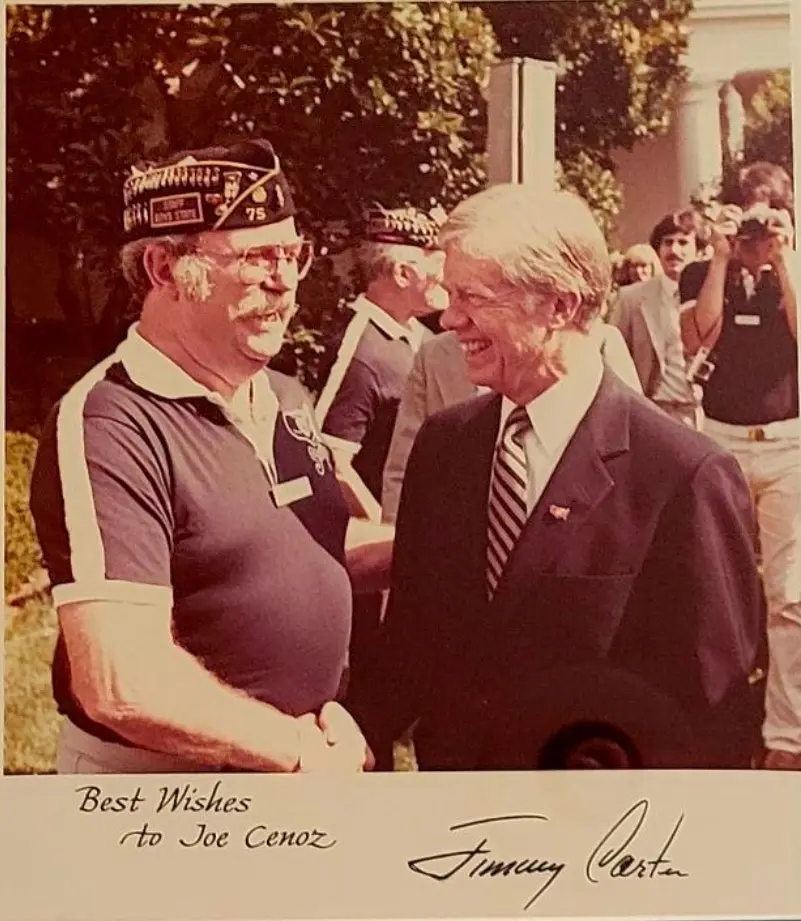 A sepia-toned photo shows former President Jimmy Carter smiling while shaking hands with a man in a military-style cap and…