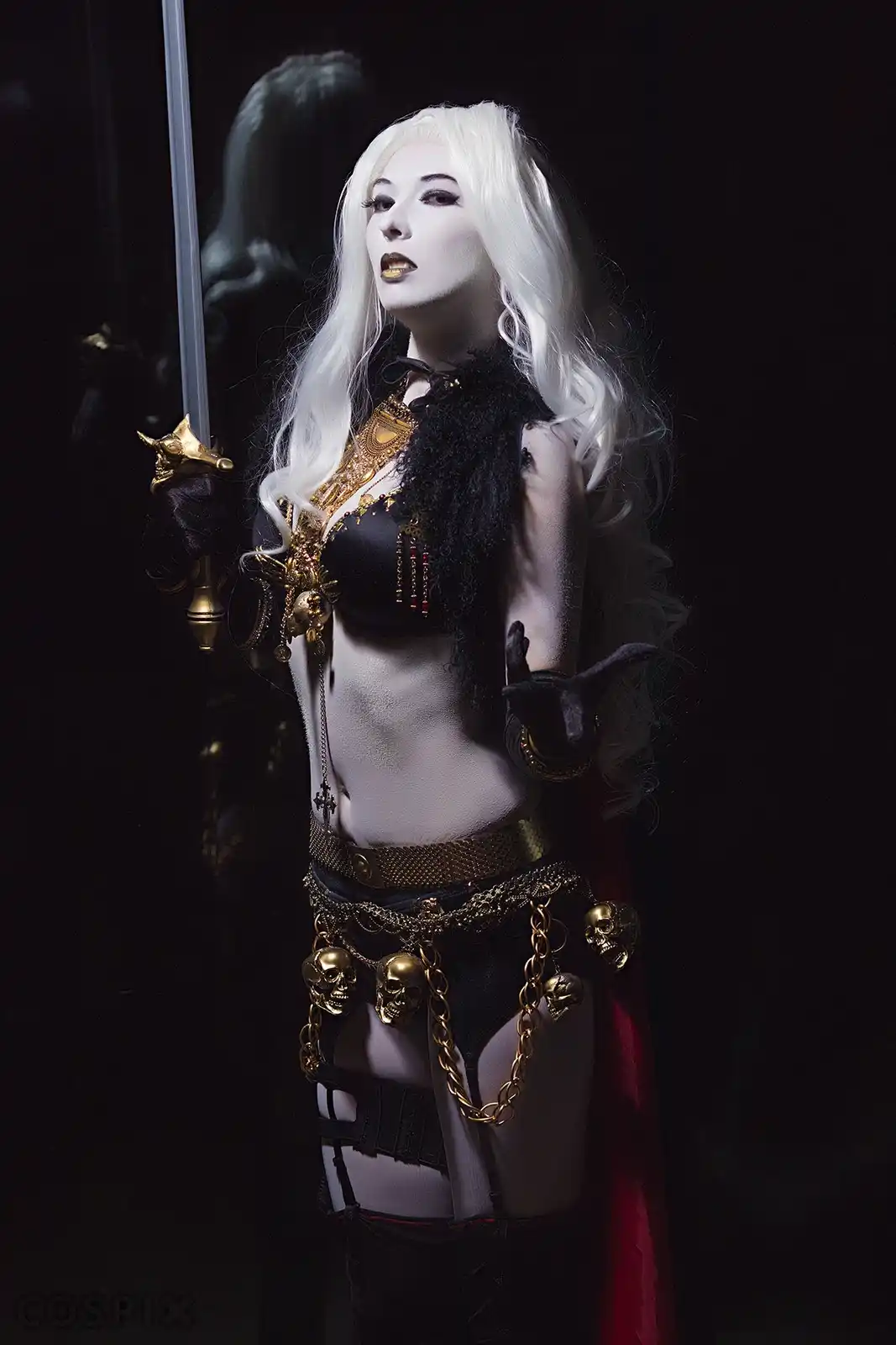 A person with long white hair and pale makeup poses in a dark, gothic costume featuring black lingerie, gold chains, skull…