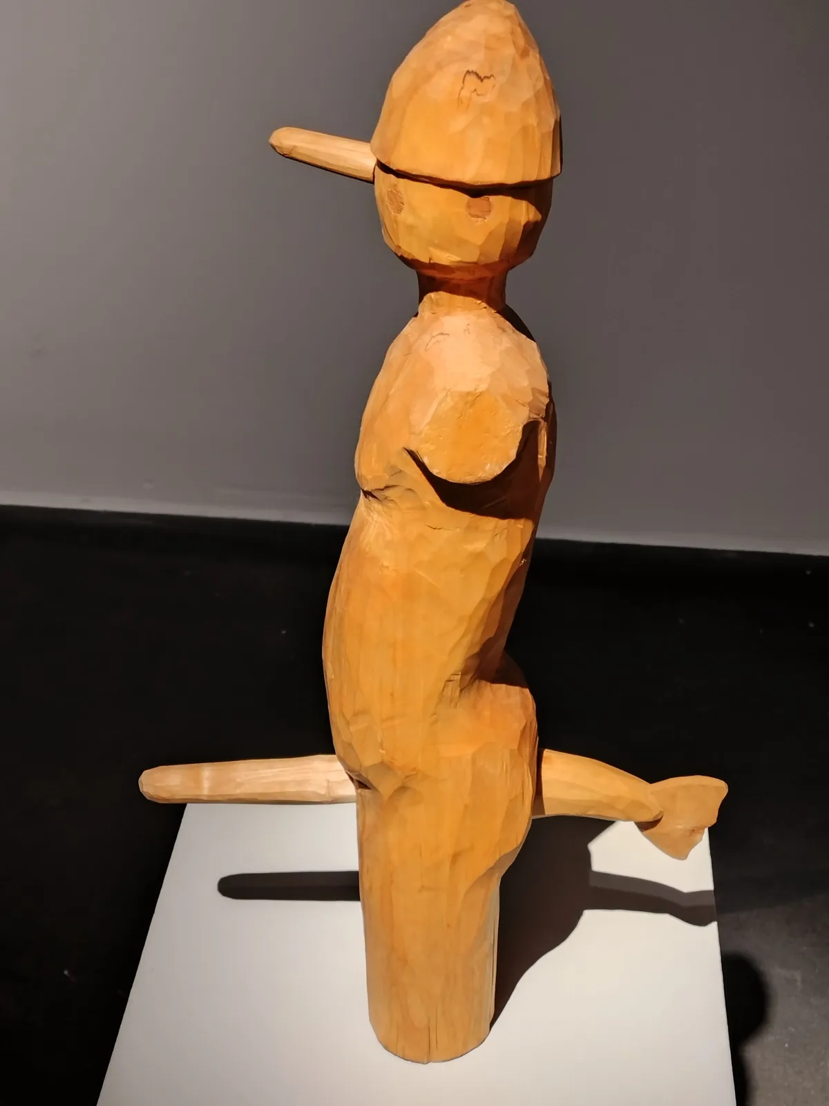 A wooden sculpture of a humanoid figure with a long nose, resembling Pinocchio, is shown from the side. The figure has a…