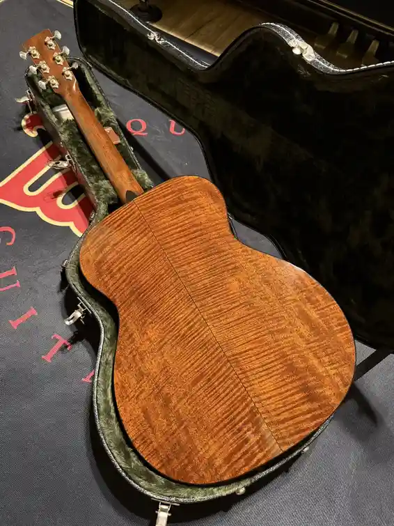 a close-up photo of an acoustic guitar with a highly figured, flame maple back resting inside its open black case. The…