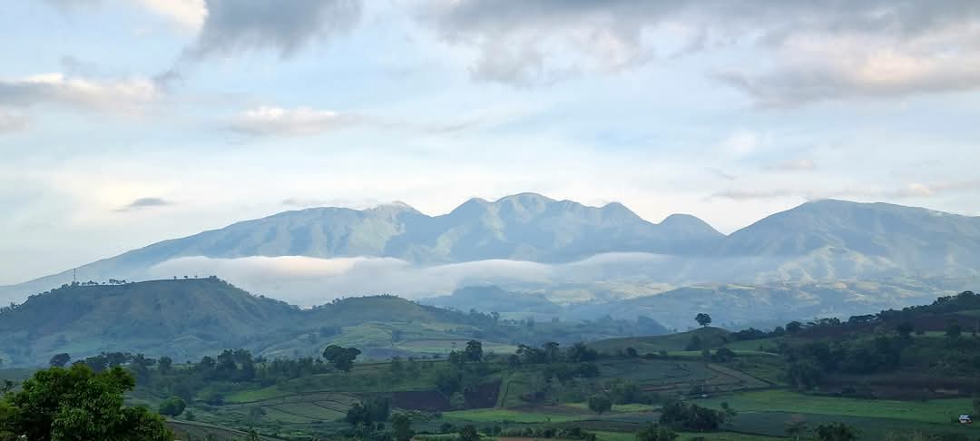 A scenic landscape photo of rolling green hills and distant mountains partially obscured by low-lying clouds or fog under a…