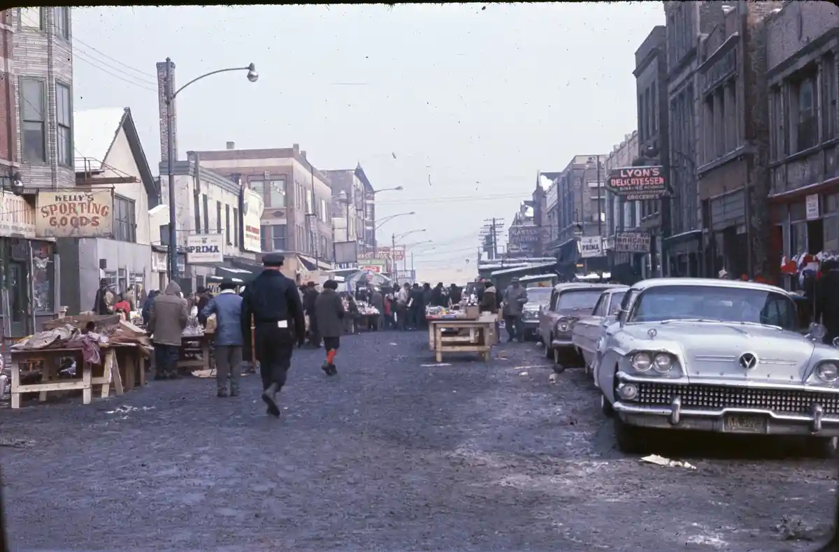 A vintage photograph shows a bustling street market with people walking among wooden vendor tables, parked cars including a…
