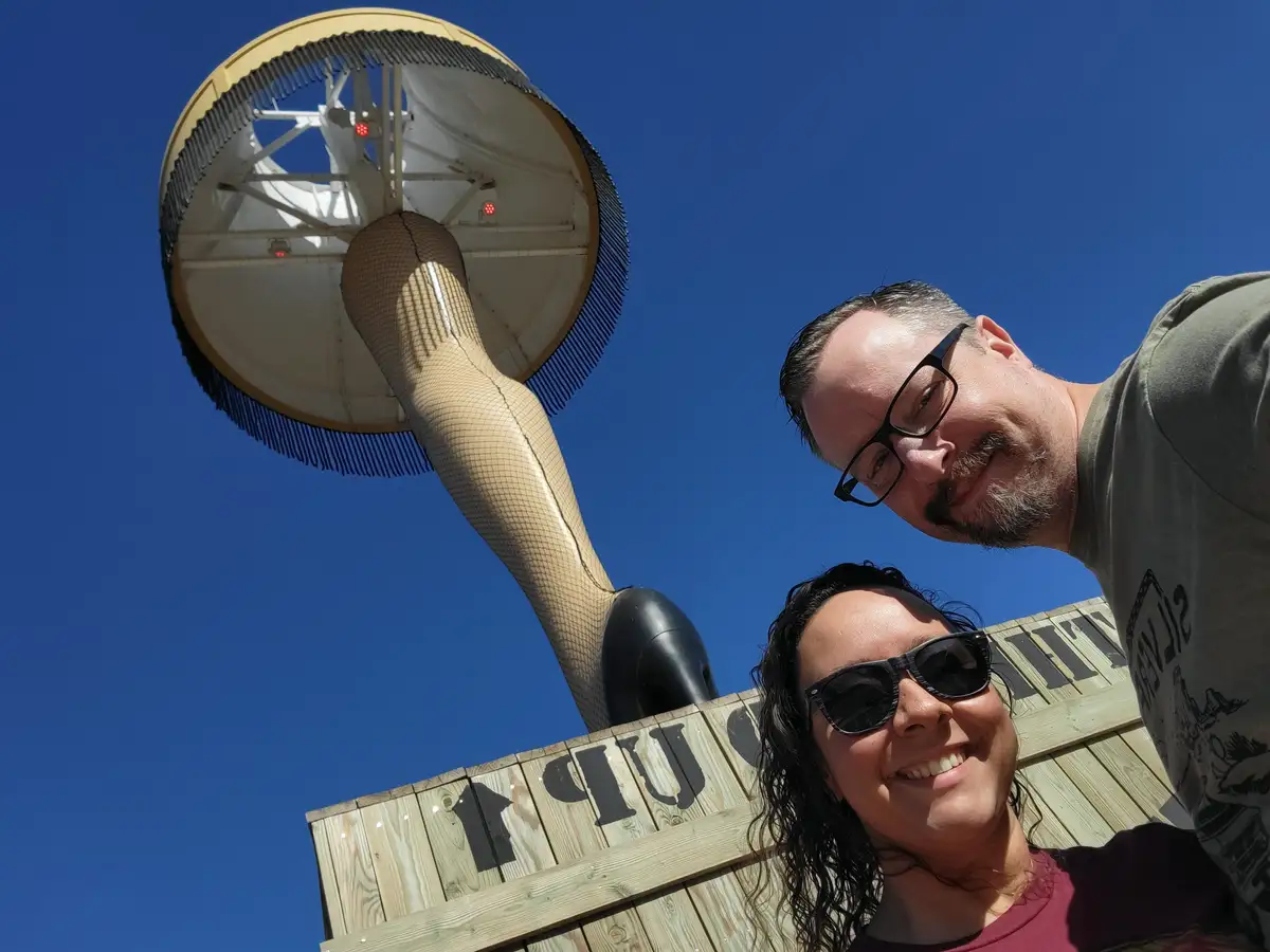 A low-angle selfie shows a smiling man and woman posing in front of a large, stylized sculpture of a woman's foot wearing a…