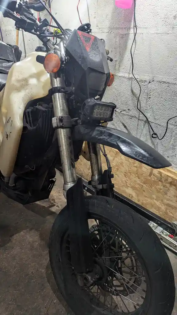 A close-up photo of a heavily modified, dirty motorcycle in a garage, featuring a custom black front fender, a large LED…