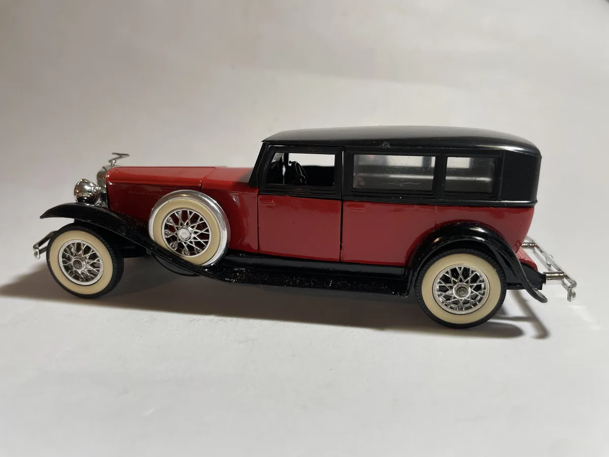 A red and black vintage-style toy car model with whitewall tires and chrome details is photographed against a plain white…