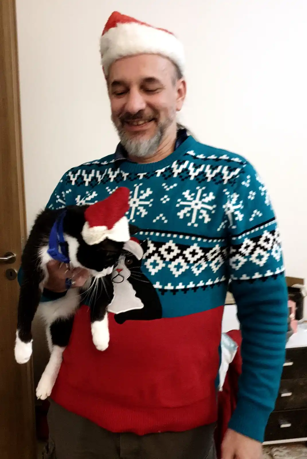 A man with a beard wearing a red and white santa hat and a festive blue and red sweater with snowflake patterns holds a…