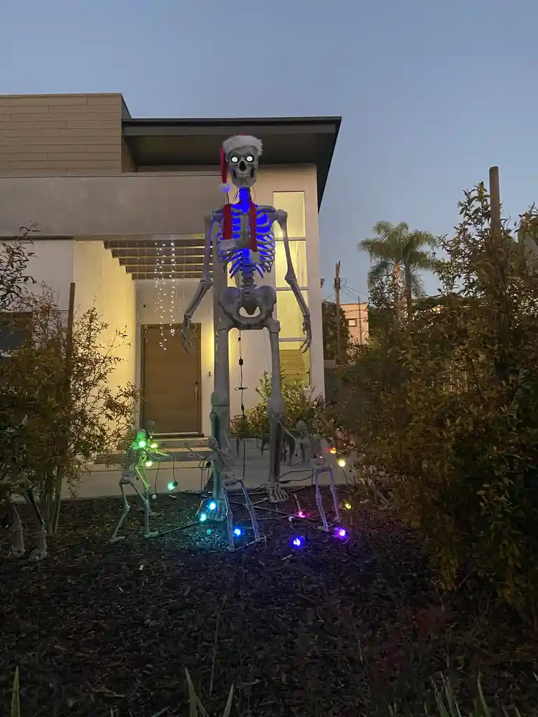 A large, illuminated skeleton wearing a Santa hat and red scarf stands in a yard at dusk, surrounded by smaller skeletons…