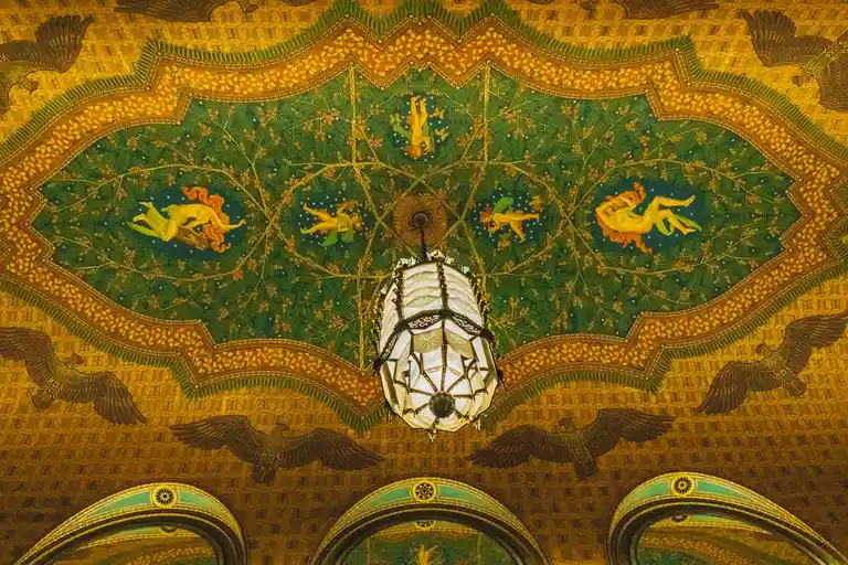 An ornate, golden ceiling with intricate patterns, painted figures of winged humans and birds, and a central chandelier. The…