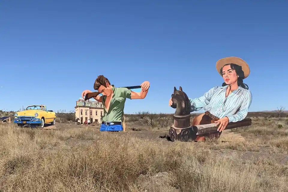 A desert landscape features giant painted cutouts of a man in a green shirt and blue pants holding a rifle, and a woman in a…