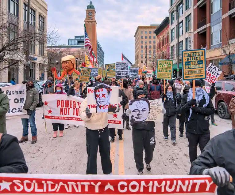A large protest march in a city street features demonstrators holding signs with political messages. Visible signs include…