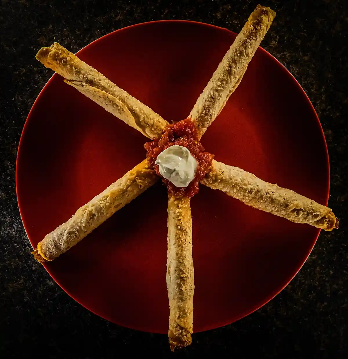 five golden-brown taquitos are arranged radially on a red plate to form a star shape, with a dollop of sour cream atop a…