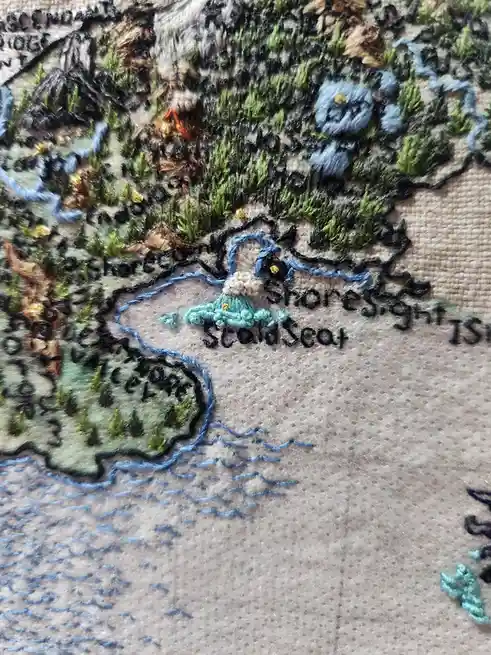 A close-up of an embroidered fantasy map showing a small island labeled 'Scald Seat' with a volcano and surrounding waters…