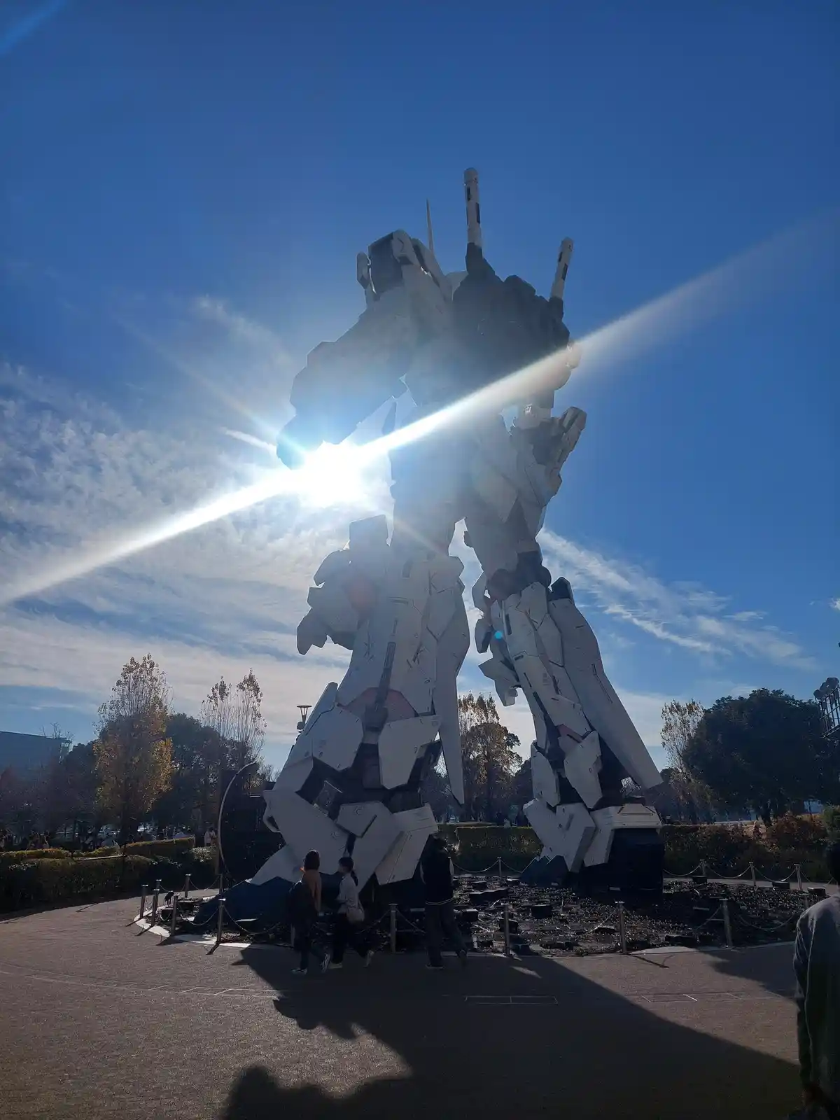 A giant white and black robot statue stands outdoors under a bright blue sky with wispy clouds, with the sun positioned…