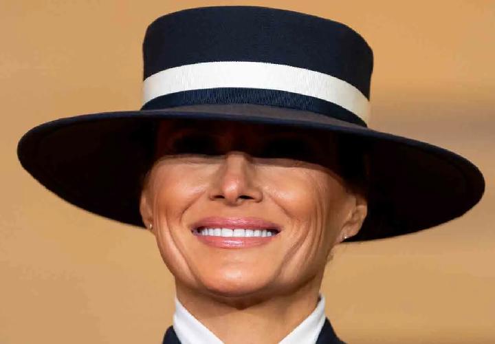 a close-up photo of a smiling woman wearing a black wide-brimmed hat with a white band, which obscures her eyes. The…