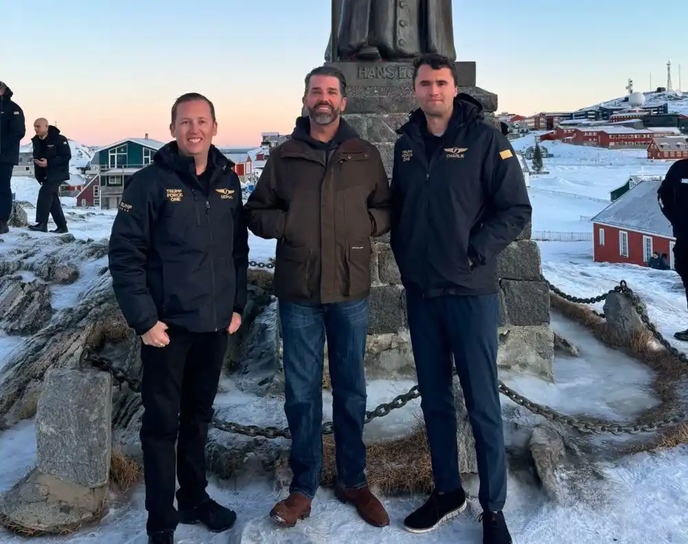 Three men stand in front of a statue in a snowy, coastal town at dusk. The man on the left wears a black jacket with 'TRUMP…