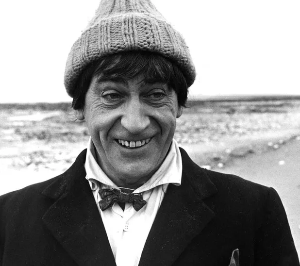 a black and white photo of a smiling man wearing a knit cap, bow tie, and coat, standing outdoors. The overlaid text reads…