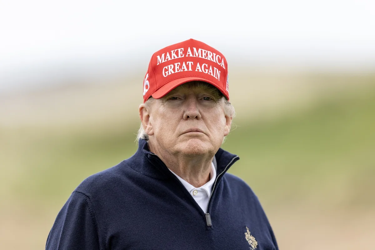 A man wearing a red 'MAKE AMERICA GREAT AGAIN' cap and a navy blue zip-up sweater stands with a serious, unamused expression…