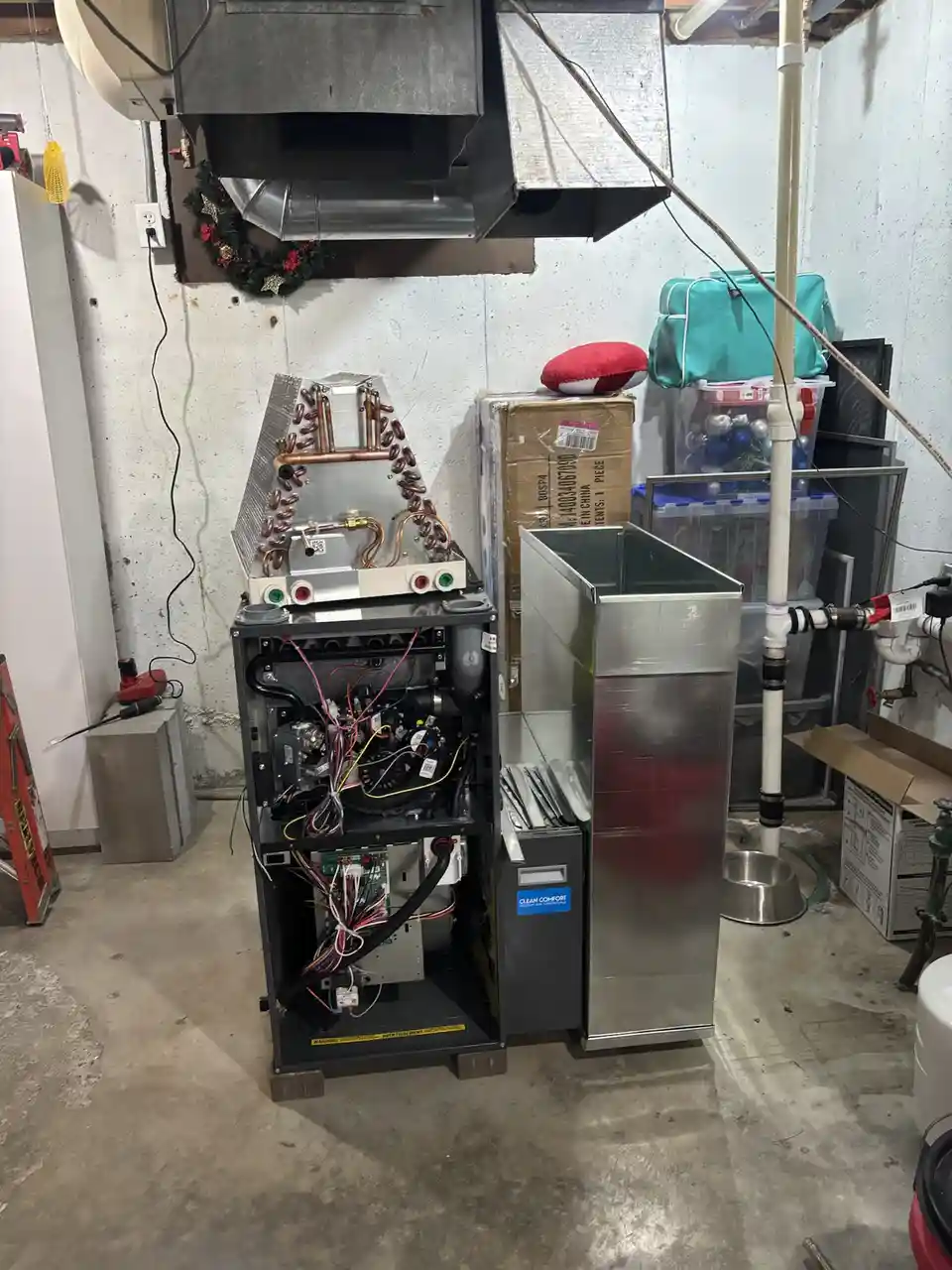 A disassembled furnace unit sits in a cluttered basement, its internal wiring exposed and copper coils visible. The…