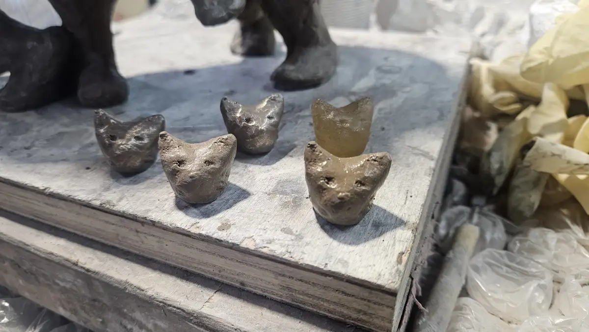 Four small, crudely shaped cat-head figurines made of clay or metal sit on a weathered wooden surface, with one appearing…