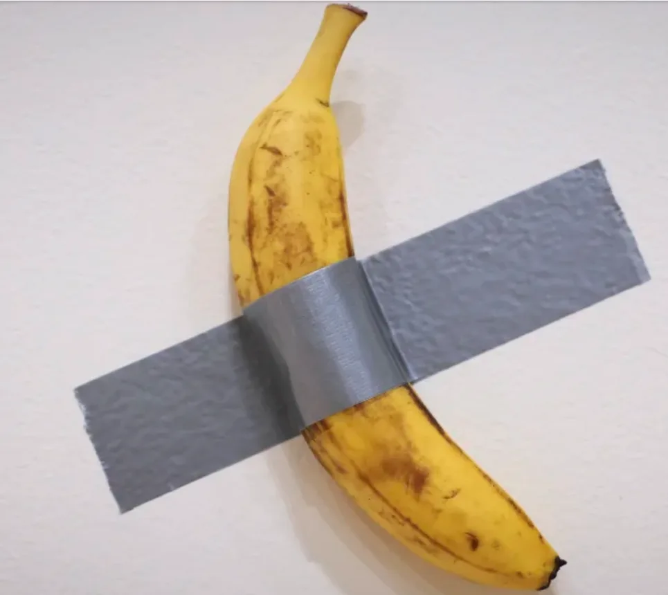 A ripe yellow banana with brown spots is taped to a plain white wall with two strips of gray duct tape forming a cross shape…