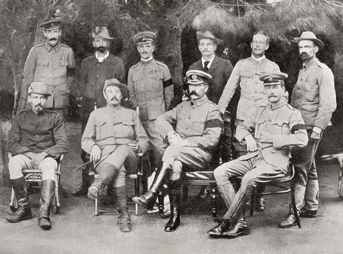 a vintage black-and-white group photo of nine men in early 20th-century military uniforms, some seated and some standing…