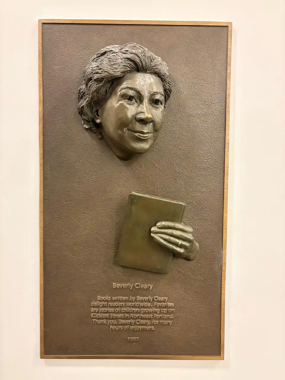 A bronze plaque mounted on a wall features a relief sculpture of Beverly Cleary's smiling face and a hand holding a book…
