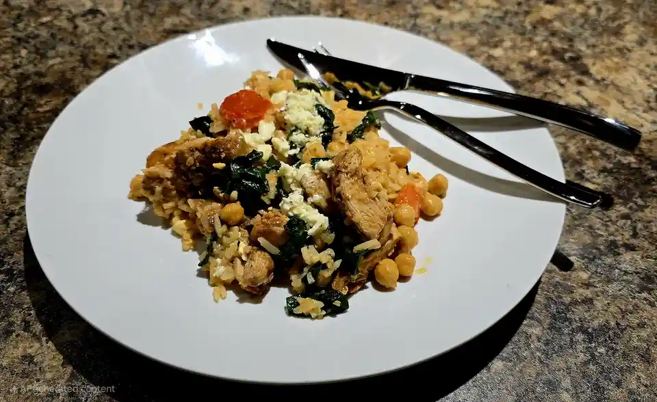 A white plate holds a colorful meal of rice, chickpeas, spinach, chicken pieces, and crumbled feta cheese, with a knife and…