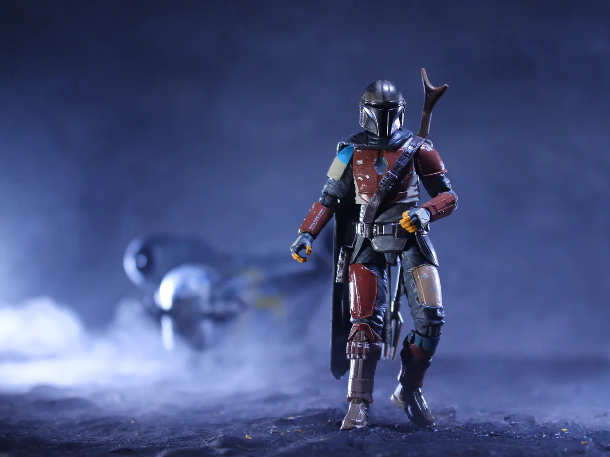 A toy action figure of The Mandalorian stands in a dramatic pose on a dark, smoky surface, facing away from the camera. In…