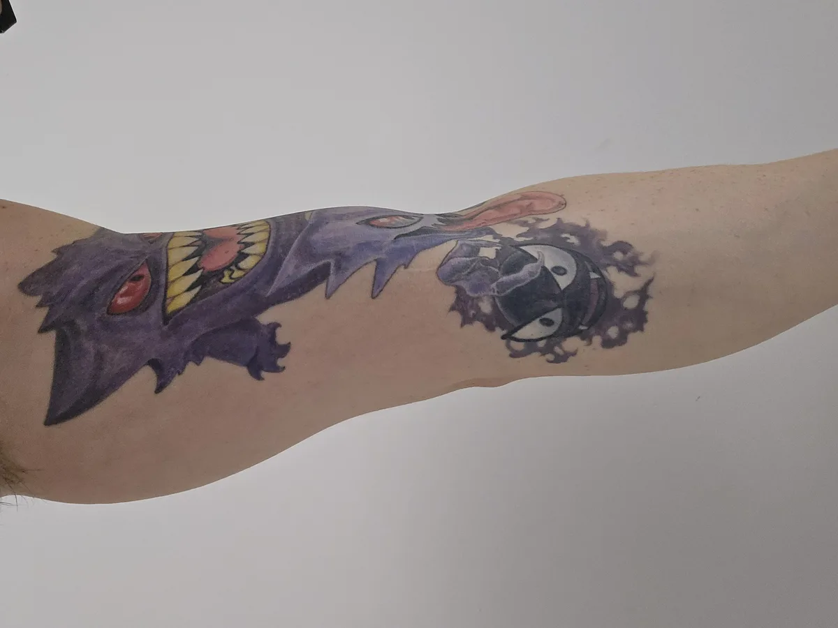 A person's forearm displays a colorful tattoo of the Pokémon Gengar, depicted with its mouth wide open as if eating another…