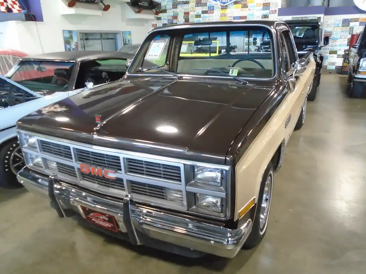 A brown and beige 1980s GMC pickup truck is displayed in a car dealership showroom, with a price tag of $24,995 visible on…