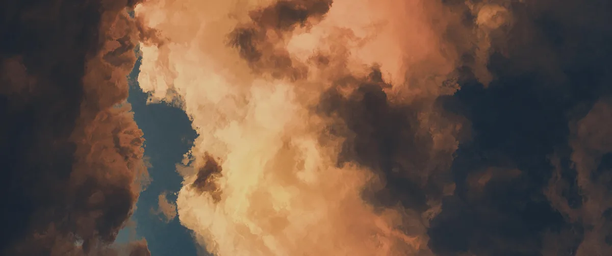 The image shows a dramatic sky filled with thick, textured clouds in shades of orange, brown, and dark blue, with a narrow…
