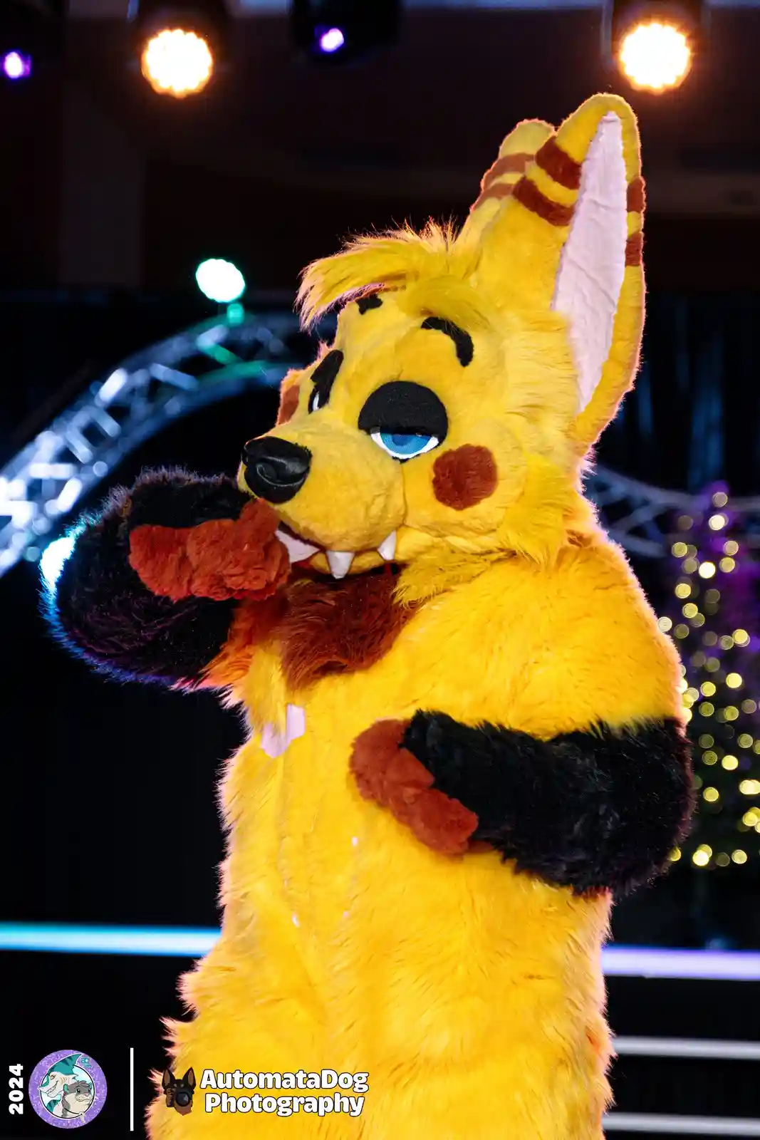 A person in a yellow fursuit resembling a fox or dog with black markings and blue eyes is posing with one hand over their…
