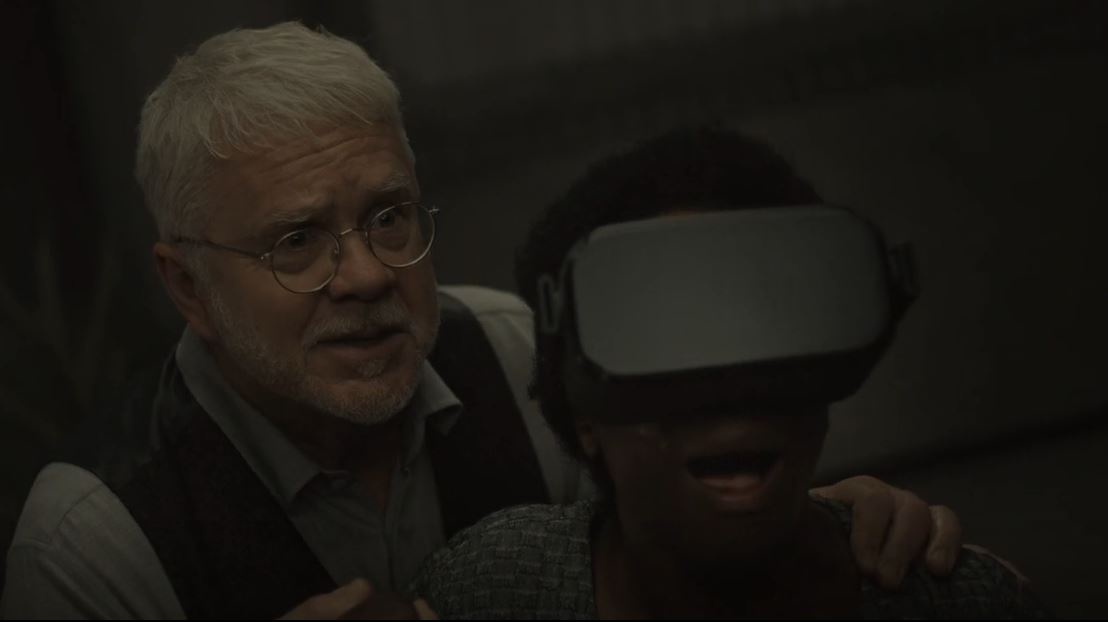 An older man with glasses and a vest has his arm around a younger person wearing a VR headset, who is screaming in apparent…