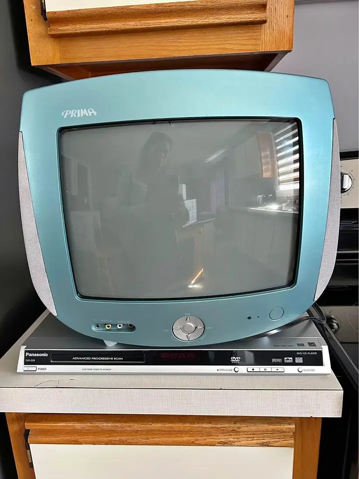 A light blue, vintage Panasonic Prima CRT television sits on top of a silver Panasonic DVD player, both placed on a wooden…